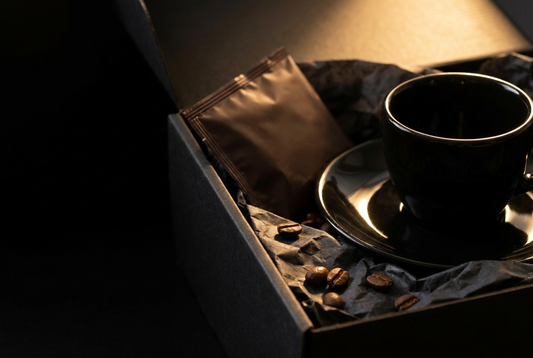 Close view of a black porcelain espresso cup and saucer in charcoal tissue inside an open black gift box, beside a sealed dark coffee pack and a few roasted beans, lit by one warm raking light.